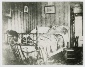 Faded photograph of an empty double bed in a small bedroom crowded with other furniture.