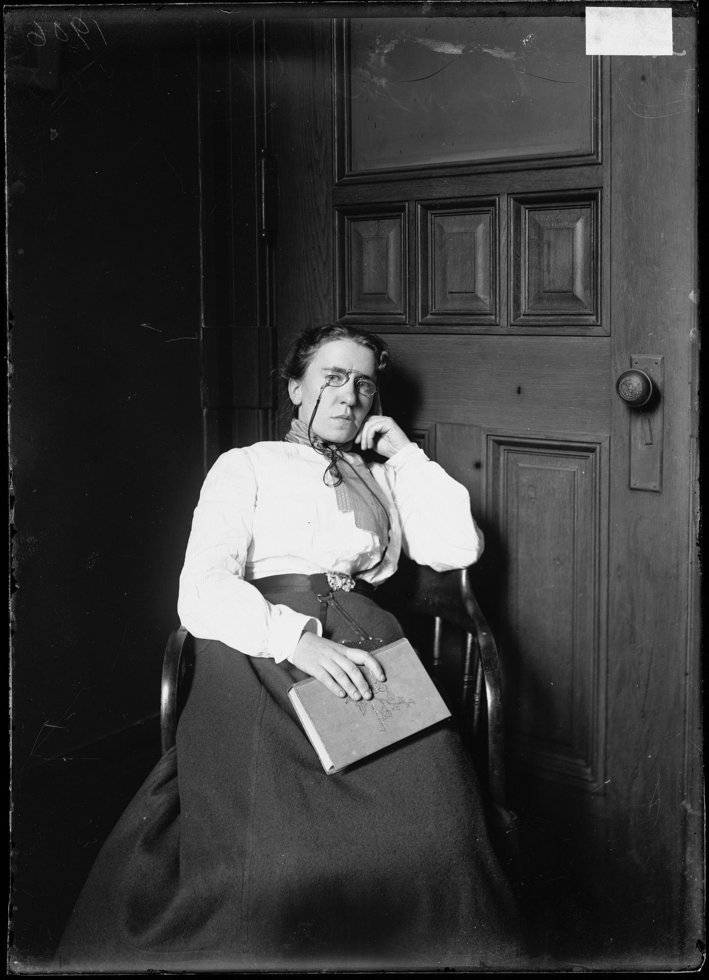 A black & white three-quarter length portrait of a woman sitting in a chair and facing the camera, in front of a wooden door.