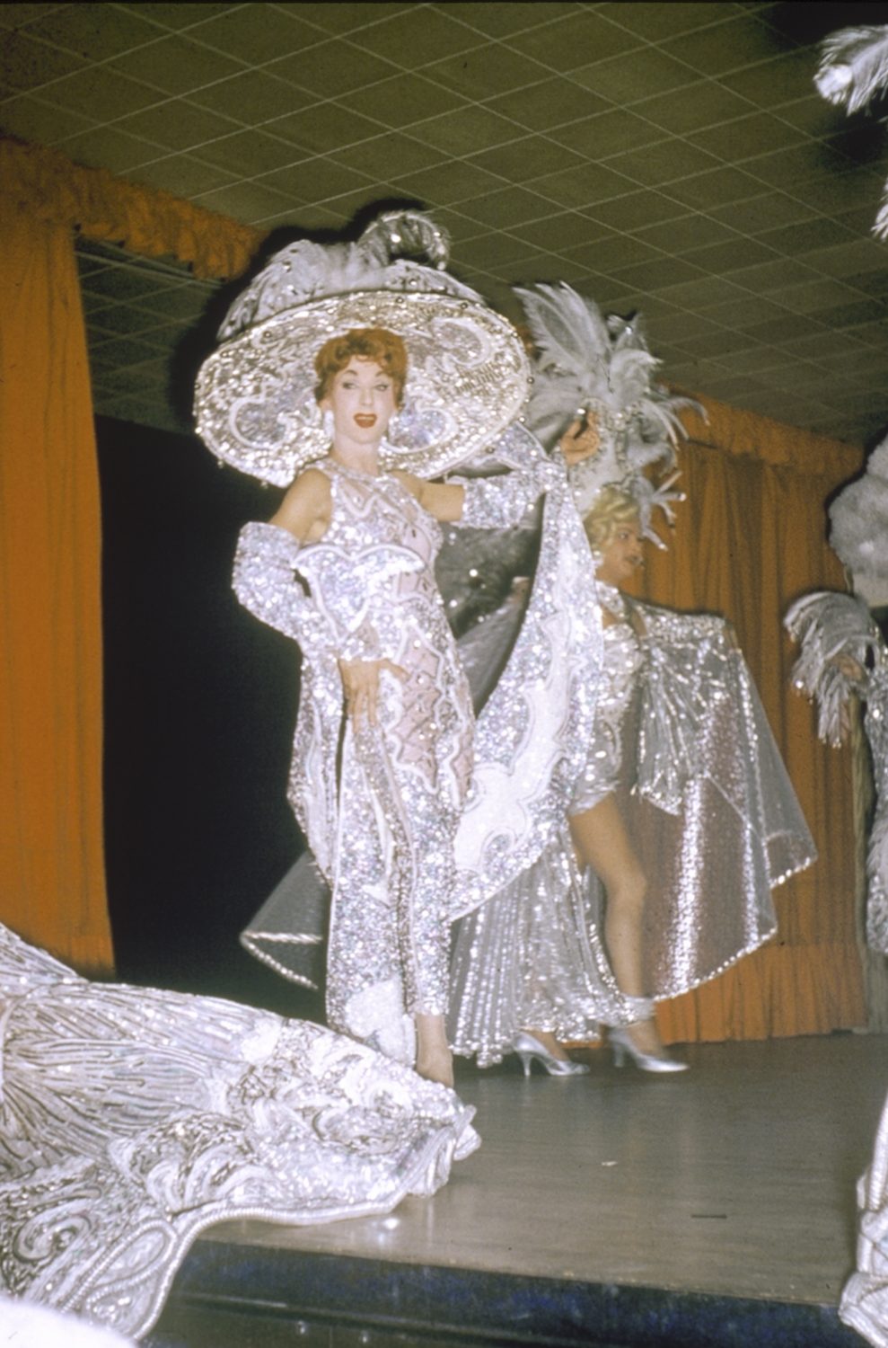 Jewel Box Revue drag show at Robert’s Show Lounge. Two female impersonators on stage – both are dressed in white gowns with large headdresses.