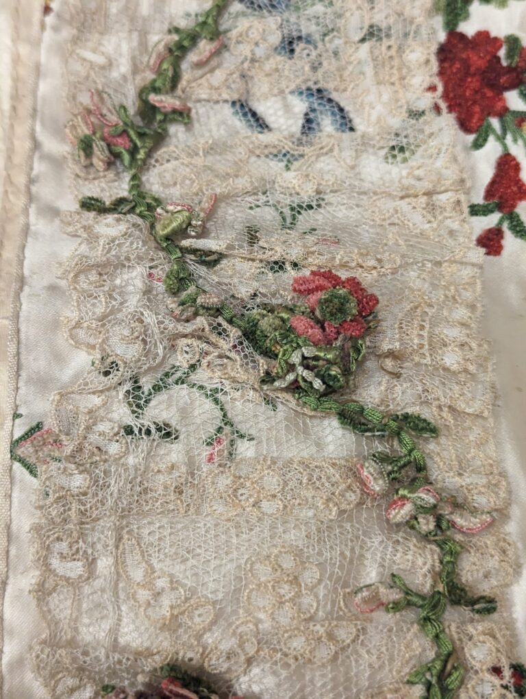 Close up view of the embroidered embellishments and lace on an 18th century robe à la francaise. On top of a strip of wide lace is a curving green vine and pink flower made out of chenille thread. 