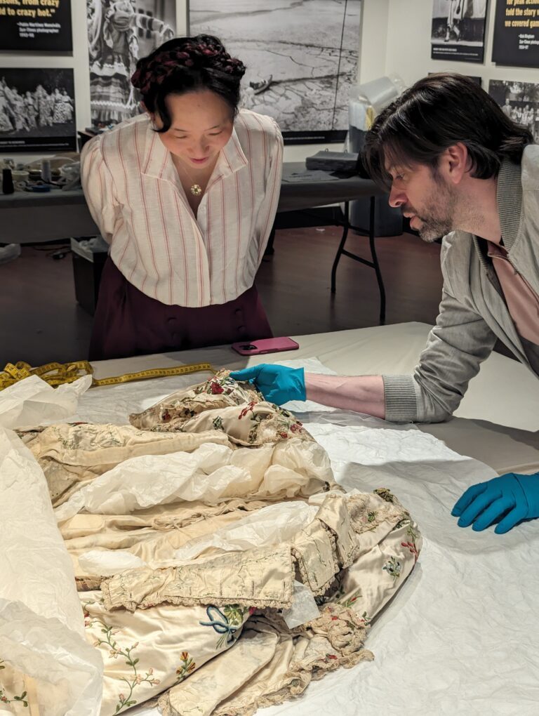 Dr. Christine Millar leans over an antique garment that is opened at the front to reveal the interior. Michael Hall holds the sleeve of the garment and wears blue gloves. 