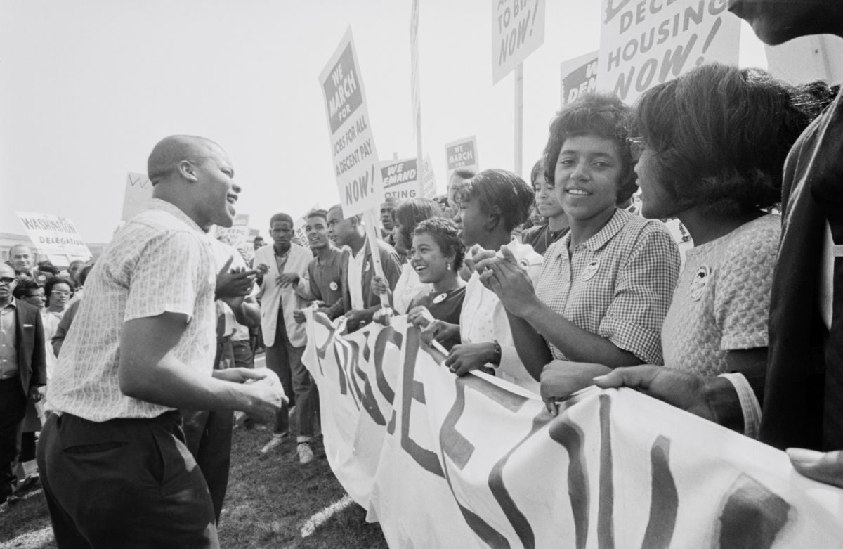 A man speaking to a smiling crowd carrying protest signs at the March on Washington for Jobs and Freedom, Washington, D.C., August 28, 1963.