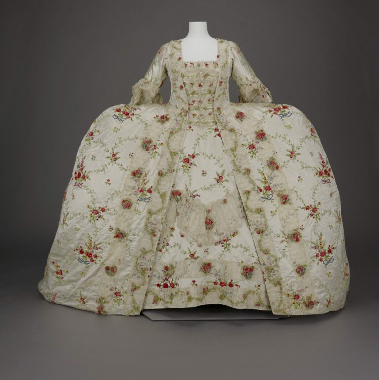 A front view of a cream colored 18th century court dress with wide panniers, and lace covering the stomacher, petticoat, and edges of the over skirt. Fabric is covered with red, yellow, and blue bunches of roses tied with red and blue bows and connected by vines. 