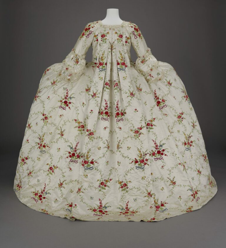 A back view of a cream colored 18th century court dress with wide panniers and lace at the sleeves. Symmetrical pleats begin at the base of the neck and reach the floor. Fabric is covered with red, yellow, and blue bunches of roses tied with red and blue bows and connected by vines. 