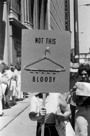 Black and white photograph of women picketing outside on a Chicago street. Image features a close-up of a sign that reads "Not this bloody" and a wire coat hanger affixed to the sign.