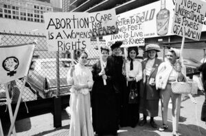 Black and white photograph of six women, some in suffrage-era costume, standing outside in front of a chain-link fence, holding signs that read "Abortion is a women's right" and "Better to die on one's feet than to live on one's knees" and "Never underestimate the powers of a women."