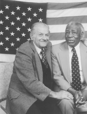 Jack Kling and Claude Lightfoot seated next to each other. Both men are in suits and ties. An American flag hangs on the wall behind them. 